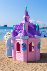 Pink plastic fairy-tale castle on beach sand. Seaside playground with loungers, umbrellas, and mountains in background.
