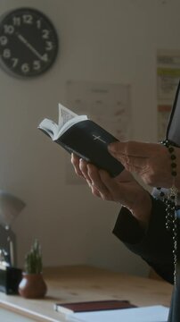Vertical cropped shot of hands of unrecognizable nun in veil holding rosary beads reading Bible in classroom