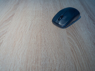 Black wireless mouse placed on the corner of a light wooden desk, ideal for office, technology, and...
