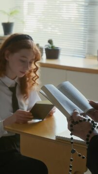 Vertical of Caucasian teen girl in school uniform sitting at desk listening carefully to unrecognizable nun in veil reading Bible out loud during catechism class in Catholic school
