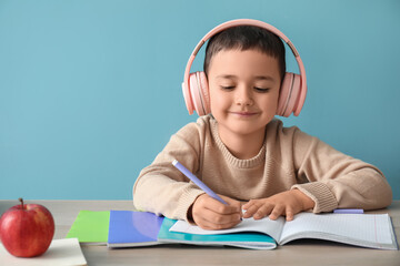 Cute little boy in headphones doing homework at table near blue wall