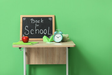 Modern school desk, stationery and blackboard with text BACK TO SCHOOL in room near green wall