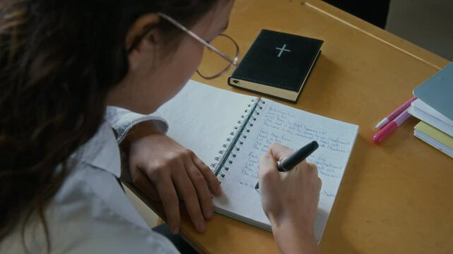 High angle shot of concentrated student girl in uniform and eyeglasses sitting at desk writing in notebook during Bible class in religious school