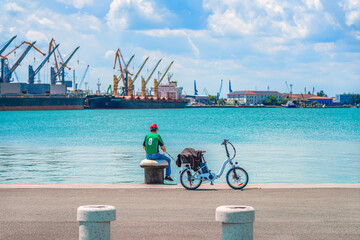 Man with fishing rod sitting on bollard near bicycle at harbor promenade, gazing over turquoise water toward industrial cranes. Concept of fishing, urban solitude, reflective outdoor moment