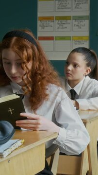 Vertical medium shot of focused teen Caucasian girl in white uniform reading Bible sitting at desk during class in Christian school
