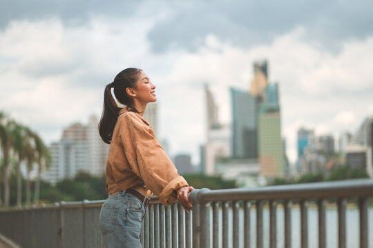 Cheerful asian woman standing on bridge in city park enjoying fresh air and nature – happy female smiling and breathing deeply in urban green space, mindful wellness lifestyle and outdoor relaxation