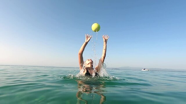 Woman joyfully splashing water while catching a ball in beach water, slow motion