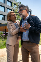 Mature businessman experiencing chest pain outdoors in business district, worried businesswoman approaching to help during possible heart emergency