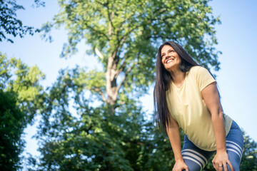 Joyful woman with serene smile resting on her knees in green park, looking up at sky and trees, feeling peace, hope and positive energy outdoors
