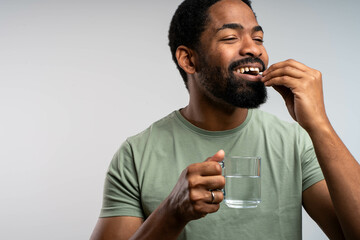 Smiling young African man taking medication with glass of water, isolated on white background, cheerful expression, health and wellness