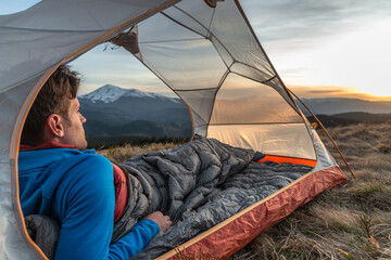 Man lying in tent with sleeping bag, watching mountain sunset view, camping in the wilderness,...