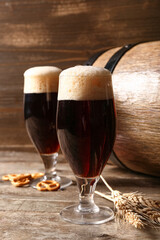 Glasses and barrel of cold dark beer with wheat and pretzels on table against wooden background