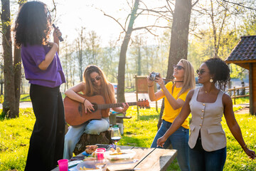 Four friends singing, playing guitar, taking photos and enjoying a picnic in the park
