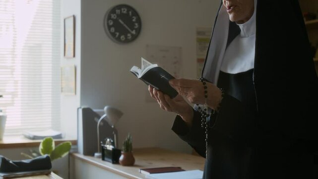 Cropped side view shot of concentrated elderly nun in veil with rosary beads in hand reading Bible out loud during catechism class in Catholic school