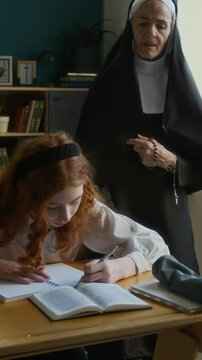 Vertical of mature Caucasian nun teacher in veil checking multiethnic student girls assignment during catechism class in religious school