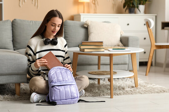 Beautiful happy female student putting books into backpack on floor at home