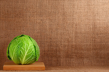 Fresh green cabbage sits on a small wooden cutting board against a burlap fabric backdrop.