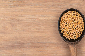 A dark wooden spoon is filled with small yellow soybeans against a textured wooden background.