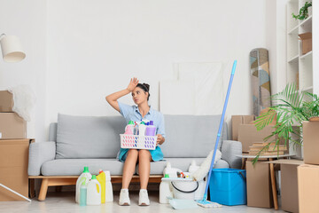 Tired female janitor with cleaning supplies sitting in room on moving day