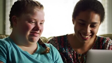 Chest up of smiling Caucasian girl with cerebral palsy speaking with Biracial girl while staying at home together during leisure time watching film on laptop