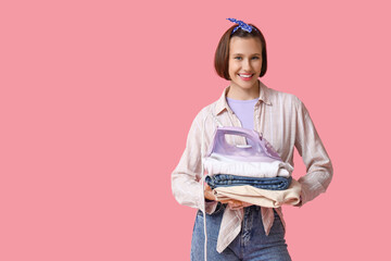 Smiling young woman with iron and stack of clothes on pink background