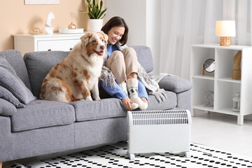 Young woman with blanket and Australian Shepherd dog sitting on sofa near radiator at home