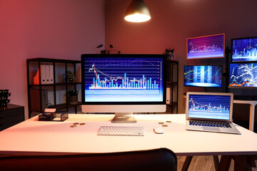 Laptop and computer monitor with stock data on trader's desk in dark office at night