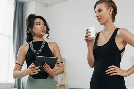 Young adult woman colleagues socializing at workplace, discussing work ideas and holding tablet and coffee, representing Gen Z business and coworker connection.