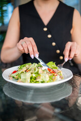 Close-up of a woman in a black dress eating a fresh tuna salad with cherry tomatoes, cucumber, and lettuce using a fork and knife at an outdoor table.