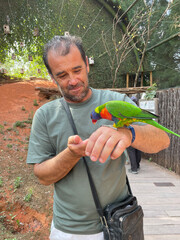 Man holding a colorful lorikeet on his hand inside a greenhouse at a wildlife reserve, highlighting friendly interaction with a tropical bird