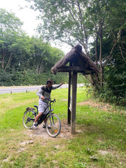 Middle-aged man on a bicycle stopping in front of an information map at the entrance of a forest, preparing for a nature ride
