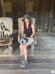 Middle-aged woman wearing safari clothes, sitting inside a traditional African hut with a wide-brimmed hat and a telephoto camera, ready for wildlife photography