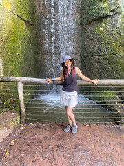 Middle-aged white woman wearing hiking clothes and a sun hat standing in front of a waterfall, enjoying nature during an outdoor adventure