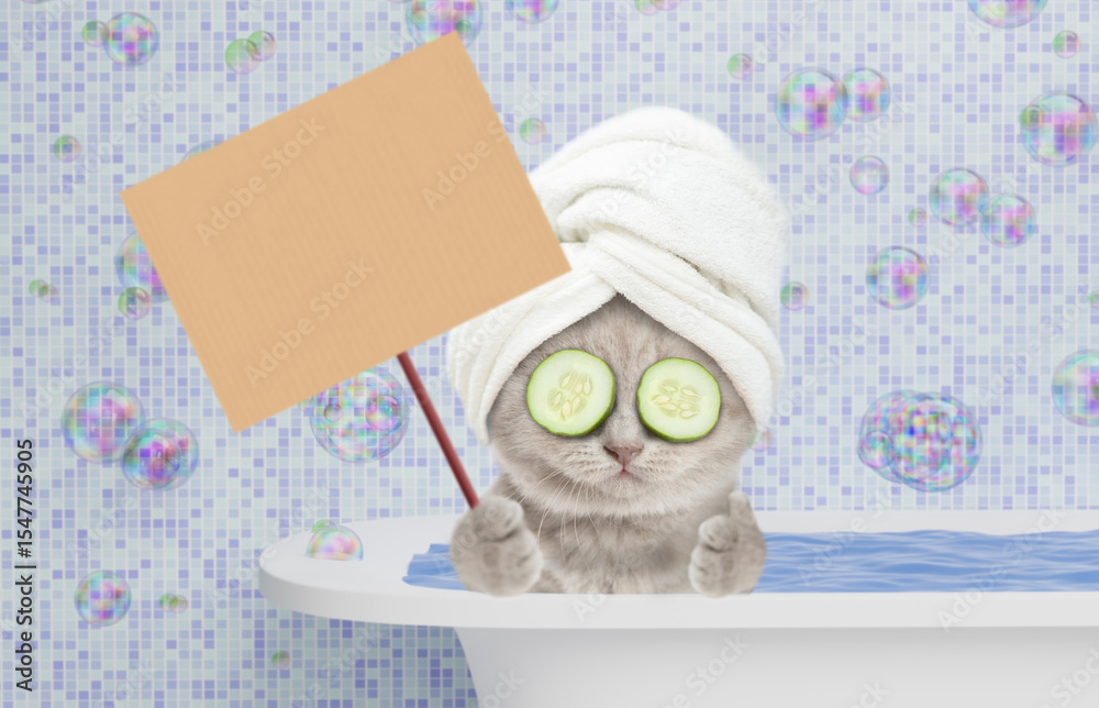 Poster Funny kitten with towel on it head and slices of cucumber on it eyes takes the bath at home and shows thumbs up gesture and blank placard on wooden stick. Empty space for text