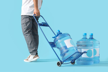 Male courier carrying trolley with bottle of clean water on blue background