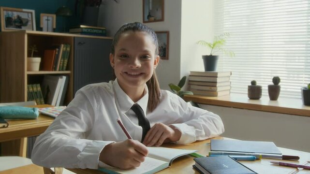 Medium portrait of concentrated teen biracial student girl in uniform writing in exercise book sitting at single desk during Bible class, then smiling at camera