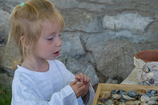 A toddler girl sitting by a table, observing sea shells during a summer DIY workshop with family.