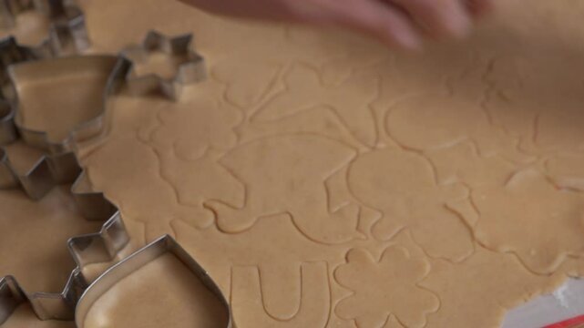 An intricate pattern of holiday cookie shapes covers a sheet of raw gingerbread dough. The detailed texture of the pastry is visible where hands have pressed in various metal cutters.