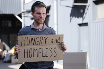 Poor man holding cardboard with text HUNGRY AND HOMELESS on street