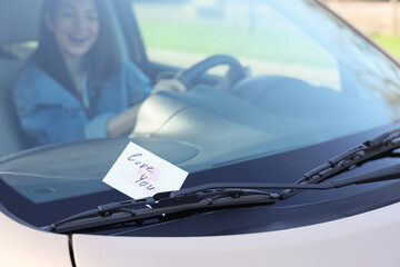 Paper with text LOVE YOU and lipstick kiss marks on windshield against woman driving car, closeup