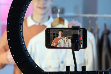 Teenage blogger with jacket recording video on screen of mobile phone in bedroom, closeup