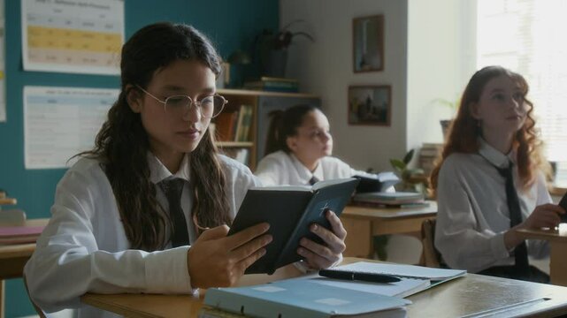 Medium slowmo of teen Caucasian and biracial girls in school uniforms sitting at single desks holding Bible and listening carefully to their teacher during religious class at school