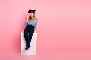 Smiling schoolgirl wearing a graduation cap holding glasses, seated on a white block against a pink...