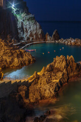 Magical night view of Manarola, Italy, with golden-lit rocky coast and calm turquoise water, creating a peaceful Mediterranean harbor atmosphere.
