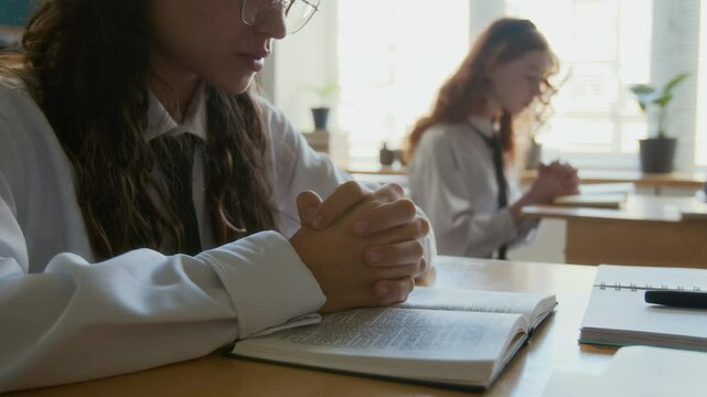 Slowmo shot of focused teen multiethnic girls in school uniforms praying during Bible class sitting at single desks in modern classroom in religious school
