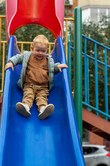 A little boy in a sweatshirt and jacket slides down a bright blue slide on a playground in the...