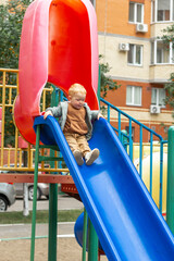 A little boy in a sweatshirt and jacket slides down a bright blue slide on a playground in the...