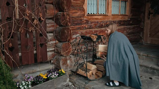 Woman wrapped in warm grey blanket, carefully carrying firewood through rustic mountain chalet interior during cold season. Blonde woman collecting and carrying wood logs through rustic mountain cabin