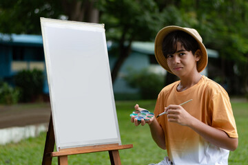 Asian teenage boy in a hat painting on a blank canvas outdoors, holding a brush and color palette, surrounded by greenery in a relaxed, creative atmosphere.