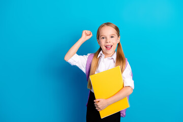Smiling schoolgirl with ponytails holding a yellow notebook against a blue background, celebrating...